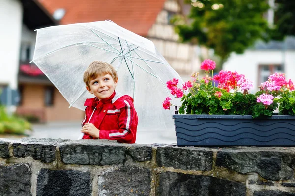 Little blond kid boy walking with big umbrella outdoors on rainy day. Portrait of cute preschool child having fun wear colorful waterproof clothes. Outdoor leisure walk on bad weather day with kids — Stock Photo, Image