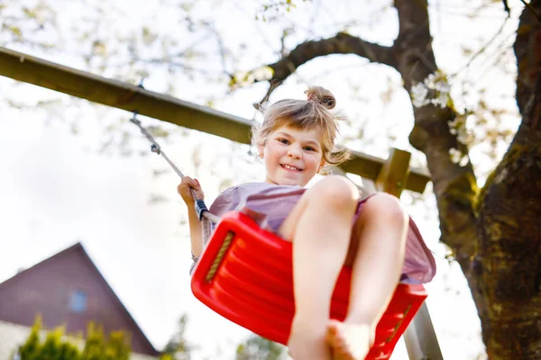 Glad liten småbarn flicka som har kul på swing i hushållsträdgården. Leende positiva friska barn svänger på solig dag. Förskoleflickan skrattar och gråter. Aktiv fritid och aktivitet utomhus. — Stockfoto