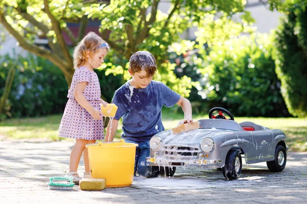 Dos niños felices lavando un gran coche de juguete viejo en el jardín de verano, al aire libre. Hermano niño y hermana pequeña niña limpiando coche con agua y jabón, divirtiéndose con salpicaduras y jugando con esponja. — Foto de Stock