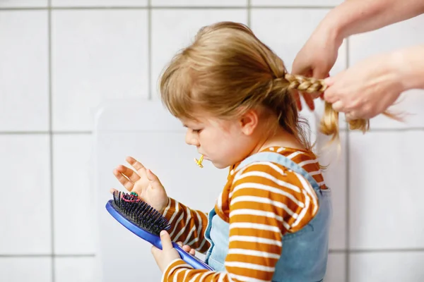 Close up photo of happy smiling toddler girl with blond hair and hands of mum brushing hairs and make plait or pigtail. Mother prepares child for kindergarten or daycare. Family morning routine. — Stock Photo, Image