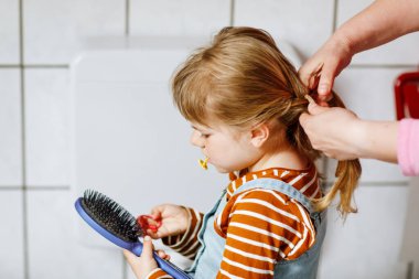 Saçlarını tarayıp örgü ya da örgü ören annenin elleri ve sarı saçlarıyla gülümseyen küçük kızın mutlu fotoğrafını çek. Anne çocuğu kreşe ya da kreşe hazırlar. Aile sabahı rutini.