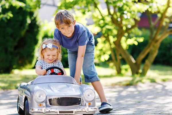 Dos niños felices jugando con un gran coche de juguete viejo en el jardín de verano, al aire libre. Chico conduciendo coche con la niña, hermana linda dentro. Riendo y sonriendo niños. Familia, infancia, concepto de estilo de vida. — Foto de Stock