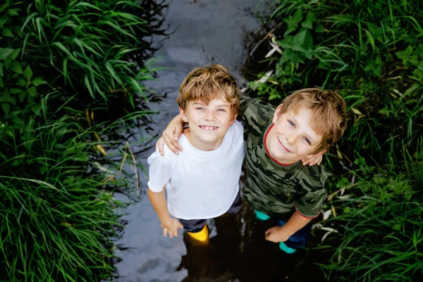 Two happy little school kids boys, funny siblings having fun together walking through water in river in gum rubber boots. Family portrait of healthy brothers and best friends — Stock Photo, Image