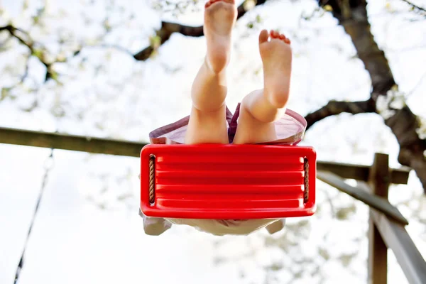 Zbliżenie stóp dziewczynki malucha bawiącej się na huśtawce w ogrodzie. Małe dziecko kołyszące się pod kwitnącymi drzewami w słoneczny dzień. Aktywny wypoczynek i aktywność na świeżym powietrzu — Zdjęcie stockowe
