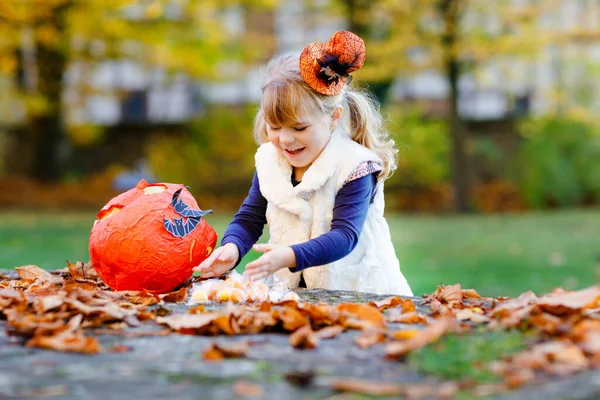Cadılar Bayramı 'nda cadı kılığına girmiş küçük bir kız. Mutlu çocuk dışarıda, turuncu komik şapkası ve tatlı lanetli balkabağı çantasıyla. Ekim 'de aile festivali sezonu. Açık hava aktivitesi — Stok fotoğraf