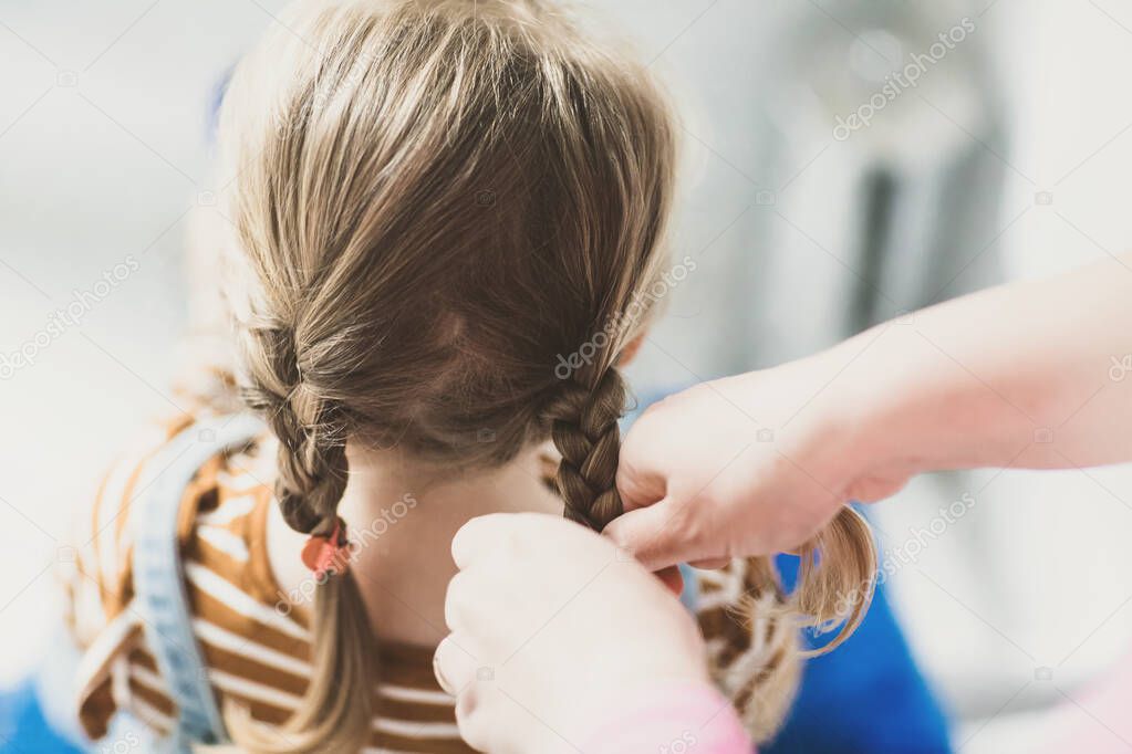 Close up photo of head of toddler girl with blond hair and hands of mum brushing hairs and make plait or pigtail. Mother prepares child for kindergarten or daycare. Family morning routine at bathroom.