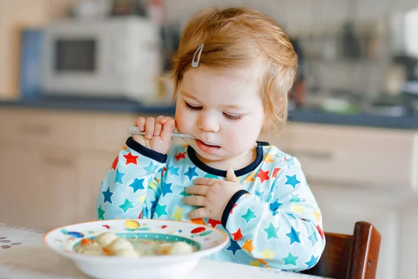 Очаровательная малышка ест с ложки овощного супа с лапшой. питание, ребенок, питание и концепция развития. Симпатичный малыш, дочь с ложкой сидит в кресле и учится есть самостоятельно. — стоковое фото