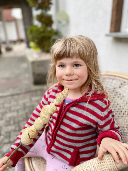 小女孩拿着用曲柄或棍子拧着的棍子面包，在火焰中烘烤着。在德国，有趣而快乐的孩子吃着受欢迎的派对和野营烧烤食品，这叫"斯托克布洛克特". — 图库照片