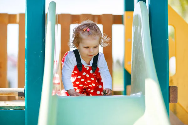 Schattig peuter meisje spelen op glijbaan op buiten speeltuin. Mooie baby in rode gom broek die plezier heeft op zonnige warme zomerdag. Kind glijdt naar beneden — Stockfoto