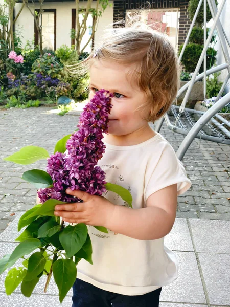 Retrato de una niña pequeña sosteniendo flores liliacas florecientes. Adorable bebé al aire libre en el cálido día de primavera. —  Fotos de Stock