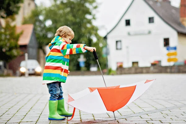 Un bambino che gioca con un ombrello quando piove. Felice bambino positivo che corre attraverso la pioggia e pozzanghere. Ragazzo con vestiti antipioggia e stivali di gomma. Attività all'aperto per bambini nella giornata del maltempo. — Foto Stock