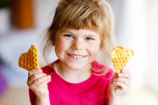 Portrét šťastné malé školačky držící čerstvě upečenou srdeční vafli. Usmívající se hladové batole se sladkou sušenkovou oplatkou. Sladké belgické vafle. — Stock fotografie