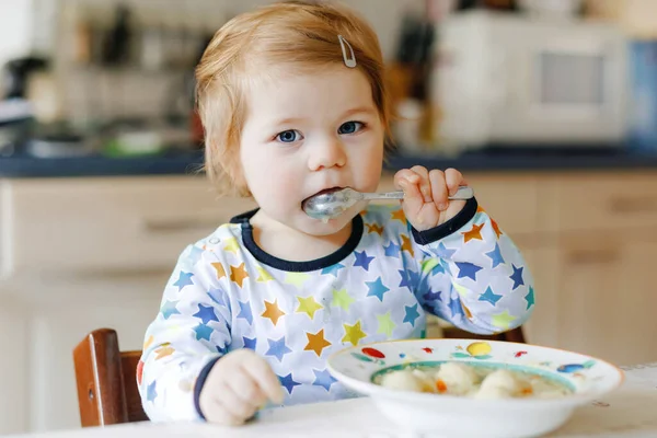 Adorabile bambina che mangia dalla zuppa di tagliatelle di verdure cucchiaio. cibo, bambino, alimentazione e sviluppo. Carino bambino, figlia con cucchiaio seduto sul seggiolone e imparare a mangiare da solo. — Foto Stock