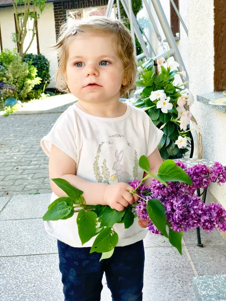 Retrato de una niña pequeña sosteniendo flores liliacas florecientes. Adorable bebé al aire libre en el cálido día de primavera. —  Fotos de Stock