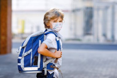 Tıbbi maskeli, sırt çantalı ya da çantalı mutlu küçük çocuk. Okul çocuğu okula gidiyormuş. Sağlıklı bir çocuk. Corona salgınından sonra karantina sonrası okula dönüş.