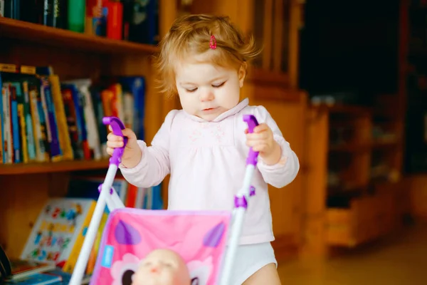 Roztomilé rozkošné holčička dělat první kroky s panenka kočár. Krásné batole dítě tlačí kočárek s hračkou doma. Šťastná dcera se učí chodit a stát — Stock fotografie