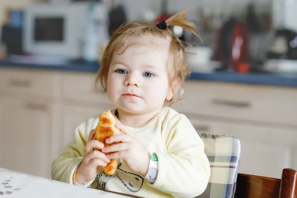 Boldog kislány, aki friss croissant-t eszik reggelire vagy ebédre. Egészséges táplálkozás gyerekeknek. Kisgyermek színes pizsamában ül a háztartási konyhában, miután reggel elaludt., — Stock Fotó