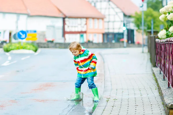 Pequeño niño jugando afuera en el día lluvioso. Feliz niño positivo corriendo y saltando a través de la lluvia y charcos. Niño con ropa de lluvia y botas de goma. Niños actividad al aire libre en el mal tiempo día —  Fotos de Stock
