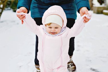 Sevimli küçük kız kışın dışarıda karda ilk adımlarını atıyor. Yürümeyi öğrenen tatlı çocuk. Anne çocuğu elinde tutuyor. Kız ve anne birlikte yürüyorlar. Mutlu aile