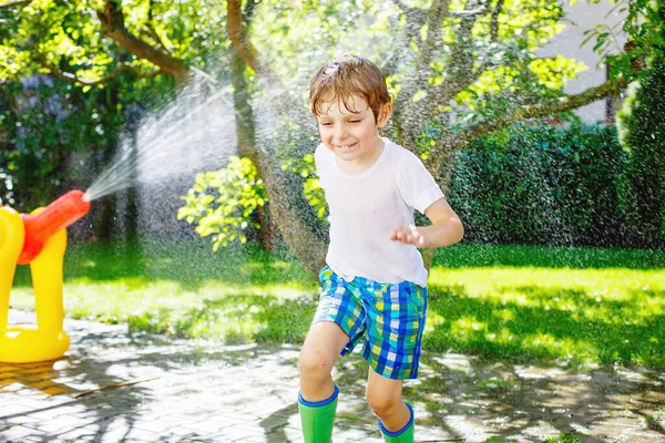 Niño divertido jugando con un aspersor de manguera de jardín en el día de verano caliente y soleado. Niño divirtiéndose con agua salpicada, gotas de lluvia. Aire libre agua de crecimiento de ocio para niños. Botas de goma para lluvia. — Foto de Stock