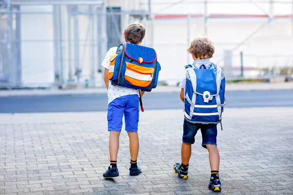 Two little kid boys with backpack or satchel. Schoolkids on the way to school. Healthy adorable children, brothers and best friends outdoors on the street leaving home. Back to school. Happy siblings.