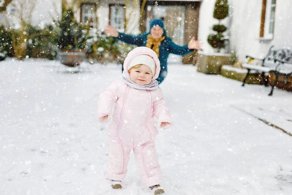 Een lief klein meisje dat 's winters de eerste stappen buiten zet. Leuke peuter die leert lopen. Gelukkige moeder op de achtergrond. Mooie familie, moeder en dochter op besneeuwde dag, wandelen door de sneeuw. — Stockfoto