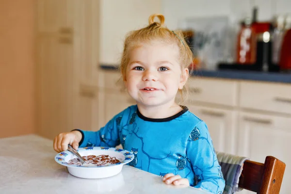 豪華な小さな幼児の女の子は朝食に牛乳で健康的なシリアルを食べています。キッチンに座って、オート、シリアルを準備するのが楽しいカラフルな服のかわいい幸せな赤ちゃん。家の中で — ストック写真