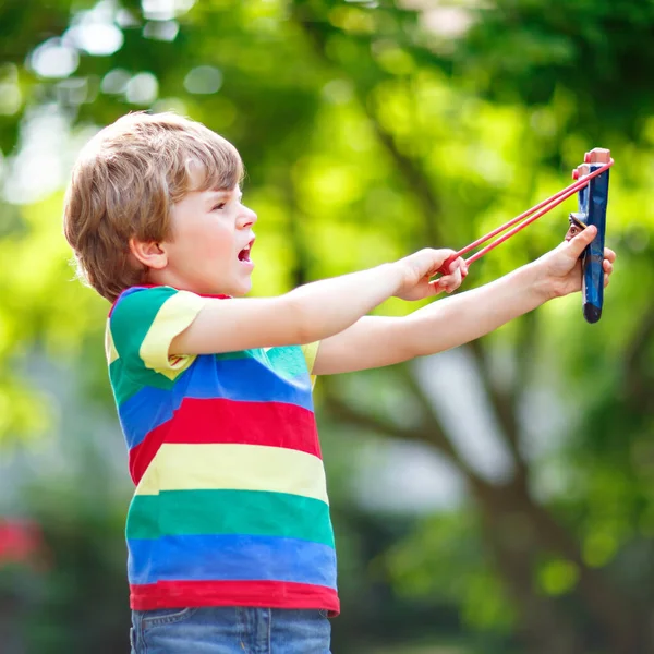 Zabawny chłopczyk strzelający drewnianą procą na zielonym tle drzewa. Dziecko bawiące się latem — Zdjęcie stockowe
