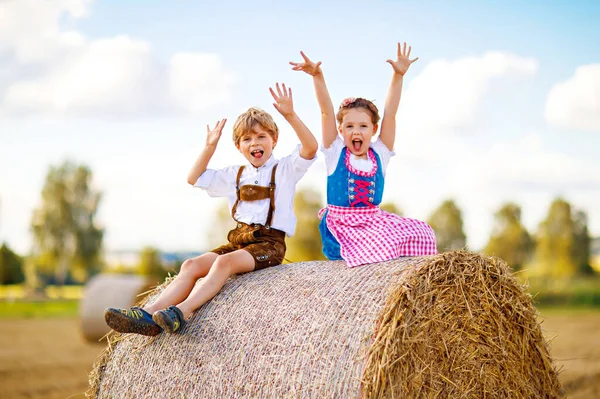 小麦の分野で伝統的なバイエルンの衣装の2人の子供。オクトーバーフェストの間に干草のベールに座っているドイツの子供たち。男の子と女の子はドイツの夏の収穫時に干し草の山で遊ぶ。親友だ. — ストック写真