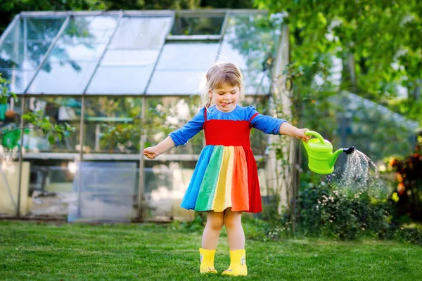 Wunderschönes kleines Mädchen in gelben Gummistiefeln und buntem Kleid, das Frühlingsblumen mit einer Wasserkanne gießt. Glückliches Kind hilft im Garten der Familie, im Freien. — Stockfoto