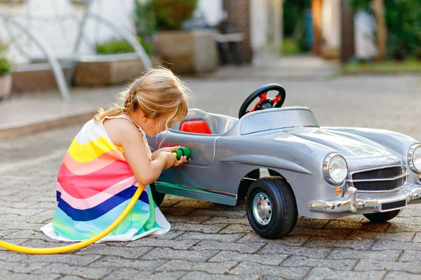 Pequeña niña jugando con un gran coche de juguete vintage y divirtiéndose al aire libre en verano. Lindo coche de repostaje infantil con agua. Chica usando manguera de jardín y llenar con gasolina, gasolinera juego de rol. —  Fotos de Stock