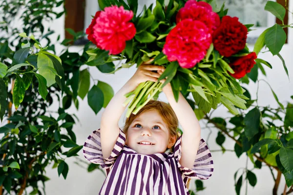 Cute adorable little toddler girl with huge bouquet of blossoming red and pink peony flowers. Portrait of smiling preschool child in domestic garden on warm spring or summer day. Summertime. — Stock Photo, Image