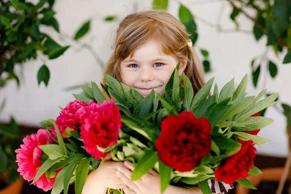 Carino adorabile bambina con un enorme bouquet di fiori di peonia rossa e rosa in fiore. Ritratto di bambino prescolastico sorridente in giardino domestico nella calda giornata primaverile o estiva. Ora legale. — Foto Stock