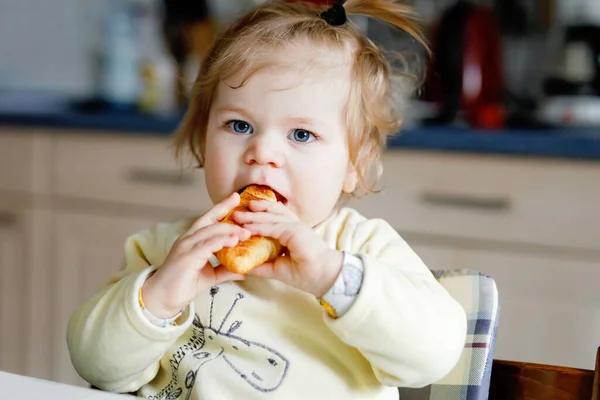 Boldog kislány, aki friss croissant-t eszik reggelire vagy ebédre. Egészséges táplálkozás gyerekeknek. Kisgyermek színes pizsamában ül a háztartási konyhában, miután reggel elaludt., — Stock Fotó