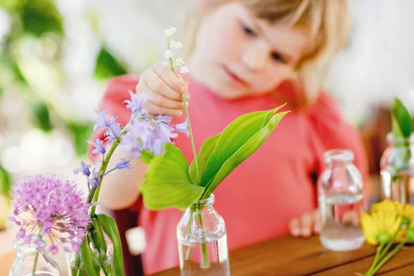 自宅で花束を作る小さな就学前の女の子。幼児の子供は水と小さなボトルにカラフルな庭の夏の花を入れます。子供のためのホームアクティビティ。虹色の花。幸せな子供. — ストック写真