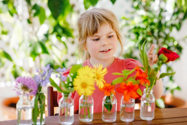 Kleine Vorschulmädchen basteln zu Hause Blumensträuße. Kleinkind legt bunte Gartensommerblumen in kleine Fläschchen mit Wasser. Aktivitäten für Kinder zu Hause. Blumen in Regenbogenfarben. Glückliches Kind. — Stockfoto