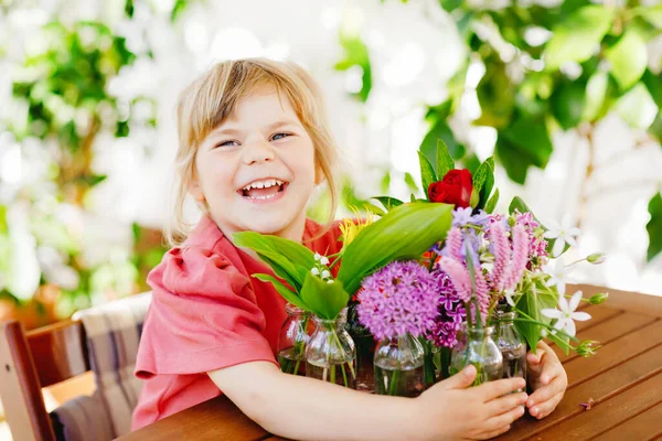 Kleines Vorschulmädchen mit Blumenstrauß zu Hause. Kleinkind legt bunte Gartensommerblumen in kleine Fläschchen mit Wasser. Aktivitäten für Kinder zu Hause. Blumen in Regenbogenfarben. Glückliches Kind. — Stockfoto