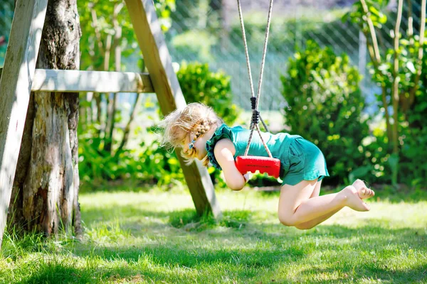 Szczęśliwa mała przedszkolanka bawiąca się na huśtawce w ogrodzie. Zdrowe dziecko huśtające się w słoneczny letni dzień. Aktywność dzieci na świeżym powietrzu, aktywny uśmiech — Zdjęcie stockowe
