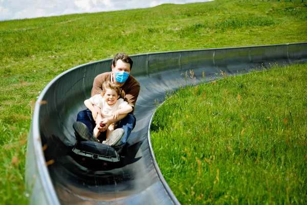 Pequeña niña y padre divirtiéndose montando trineo de verano correr trineo por una colina. Niño preescolar activo y papá con máscara médica haciendo actividad divertida otudoors. Vacaciones en familia con niños. — Foto de Stock