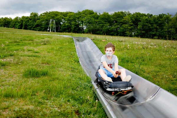 Niño de la escuela que se divierte montando trineo de verano correr trineo por una colina en Hoherodskopf, Alemania. Niño activo con máscara médica haciendo actividad divertida otudoors. Vacaciones en familia con niños. —  Fotos de Stock