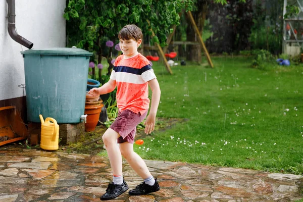 Niño de la escuela corriendo a través de fuertes lluvias de verano en el jardín. Feliz niño mojado sonriente divirtiéndose con salpicaduras y saltando en charcos. Actividad para niños en el día de lluvia. — Foto de Stock