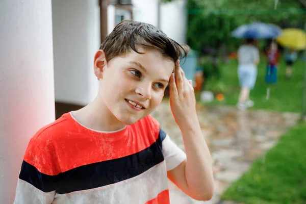 Chłopak ze szkoły biegnący przez letni deszcz w ogrodzie. Szczęśliwe uśmiechnięte mokre dziecko bawiące się chlapaniem i skakaniem w kałużach. Aktywność dla dzieci w deszczowy dzień pogodowy. — Zdjęcie stockowe