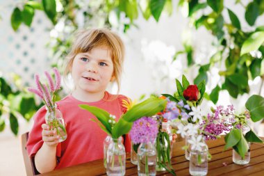 Küçük anaokulu kızı evde çiçek buketi yapıyor. Küçük çocuk renkli bahçe çiçeklerini suyla birlikte küçük şişelere koyuyor. Çocuklar için ev aktiviteleri. Gökkuşağı renginde çiçekler. Mutlu çocuk.