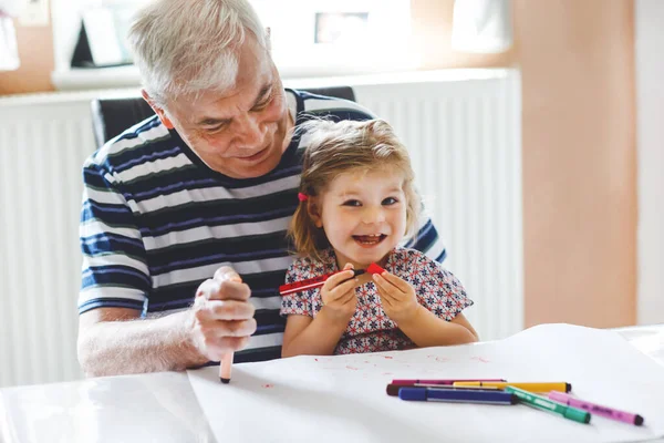 Cute little baby toddler girl and handsome senior grandfather painting with colorful pencils at home. Grandchild and man having fun together — Stock Photo, Image