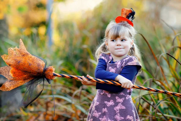 Linda niña pequeña vestida como una bruja celebra Halloween. Niño feliz al aire libre, con sombrero naranja divertido y escoba de bruja. Hermosa temporada de festivales familiares en octubre. Actividades al aire libre —  Fotos de Stock