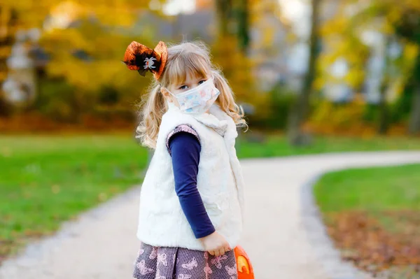 Malé batole dívka oblečená jako čarodějnice s lékařskou maskou na obličeji trik nebo léčbě na Halloween. Dítě venku, s legračním kloboukem a taškou na sladké strašení. Doba uzamčení karantény viru pandemie korony — Stock fotografie