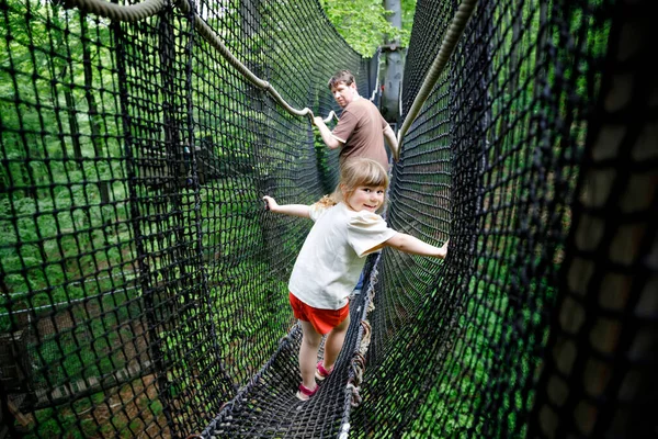 Little preschool girl and father walking on high tree-canopy trail with wooden walkway and ropeways. Happy active child and dad, young man exploring treetop path. Funny activity for families outdoors