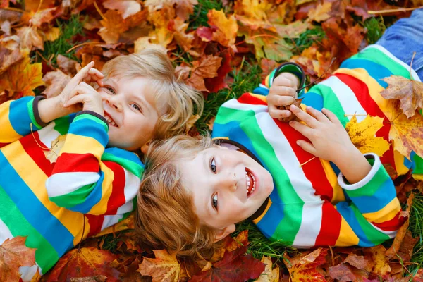 Two little twin kids boys lying in autumn leaves in colorful fashion clothing. Happy siblings having fun in autumn park on warm day. Healthy children with blond hairs and blue eyes with maple foliage. — Stock Photo, Image