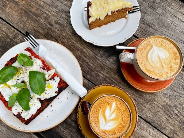 Fresh focaccia with tomato, basil and goat cottage cheese, two cups of coffee and carrot cake on wooden table. Italian food and breakfast.