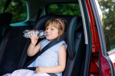 Adorable toddler girl sitting in car seat, holding bottle with water and looking out of the window on nature and traffic. Little kid traveling by car. Child safety on road. Family trip and vacations clipart